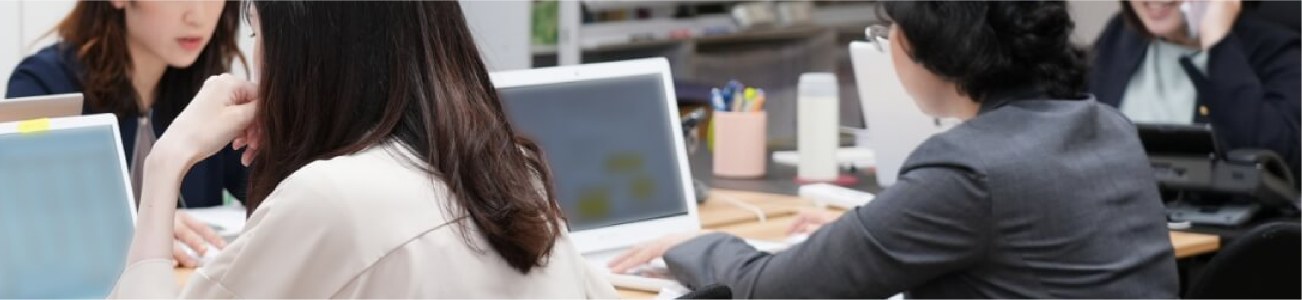 オフィスで女性スタッフがパソコン作業や電話対応をしている様子。複数人が机を囲み、業務に集中する働く姿を写した職場風景。